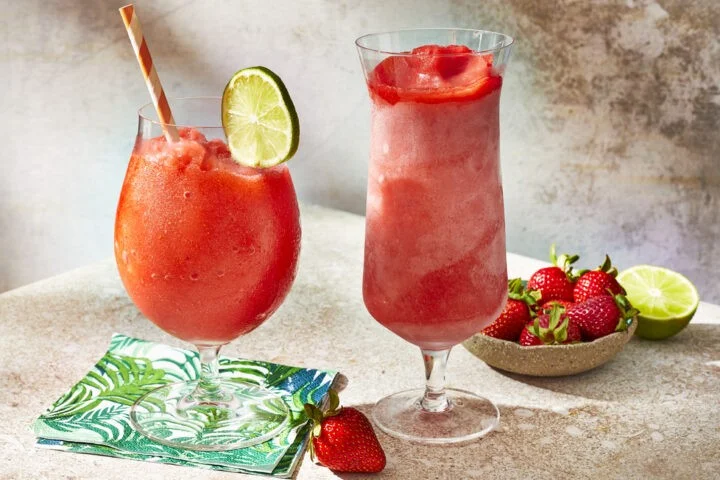 Cocktail daiquiry în două pahare înalte pe o masă cu niște căpșuni și un lime tăiat în două.